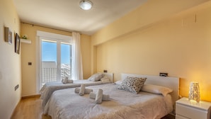 Una camera d'albergo con due letti, un balcone, un comodino bianco e una lampada da comodino.