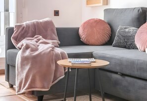 Un sofá gris con una manta rosa y almohadas, una mesita redonda con un libro y una ventana con persianas.