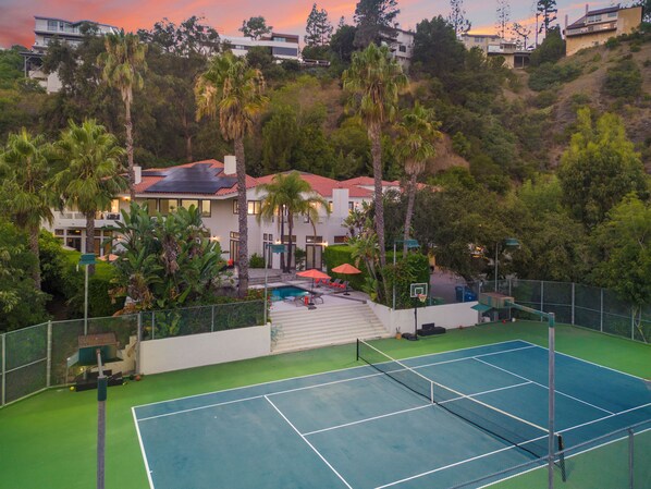 A tennis court with a net, surrounded by a fence. There are palm trees and a building with a red roof in the background.