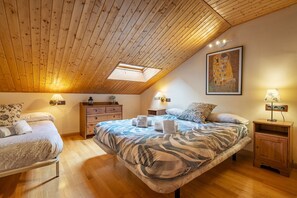 A bedroom with a sloped ceiling, a bed with a patterned bedspread, two bedside tables with lamps, a nightstand, and a framed painting on the wall.
