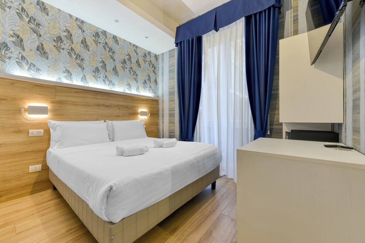 A modern hotel room with a wooden headboard, a bed with white linens, a bedside table, and a large window with blue curtains.