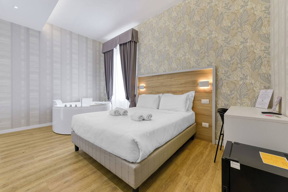 A modern hotel room with a large bed, a bedside table, a built-in desk, and a shower area.