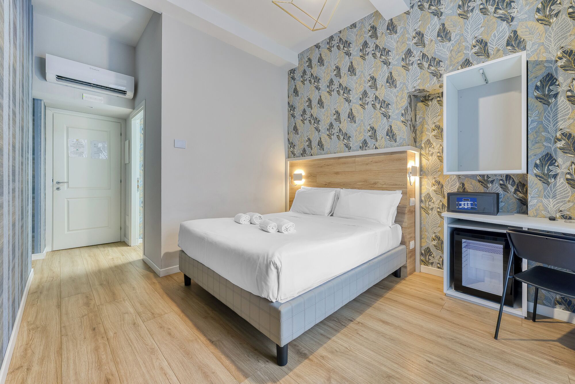 A hotel room with a bed, a desk, a mirror, and a wall with a leaf pattern.