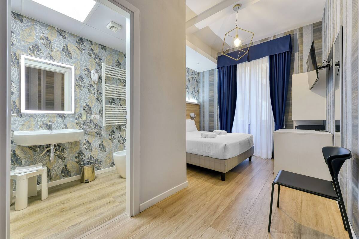 A modern hotel room with a bathroom, a bed, a desk, and a chair.