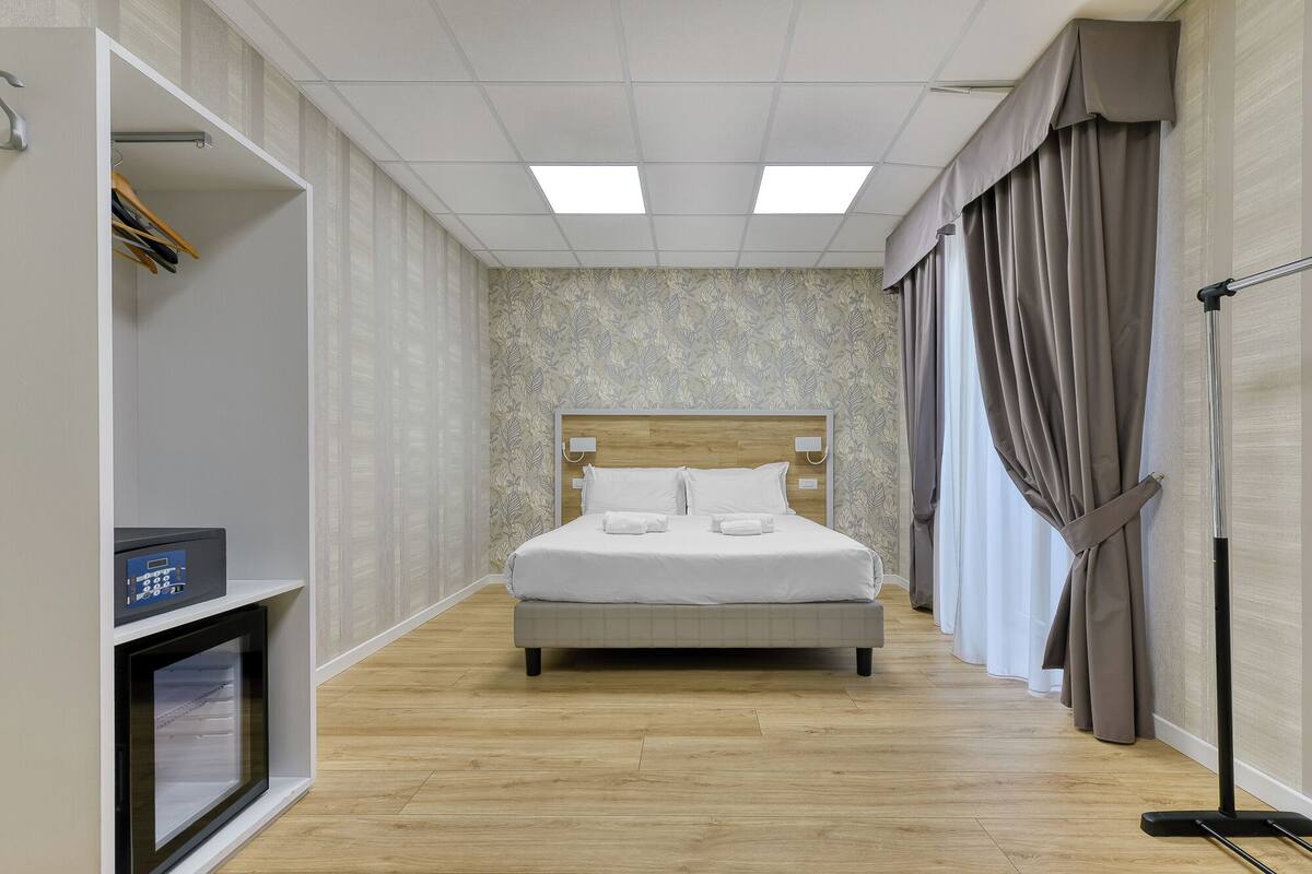 A hotel room with a bed, a desk with a computer, a wardrobe, and a window with curtains.