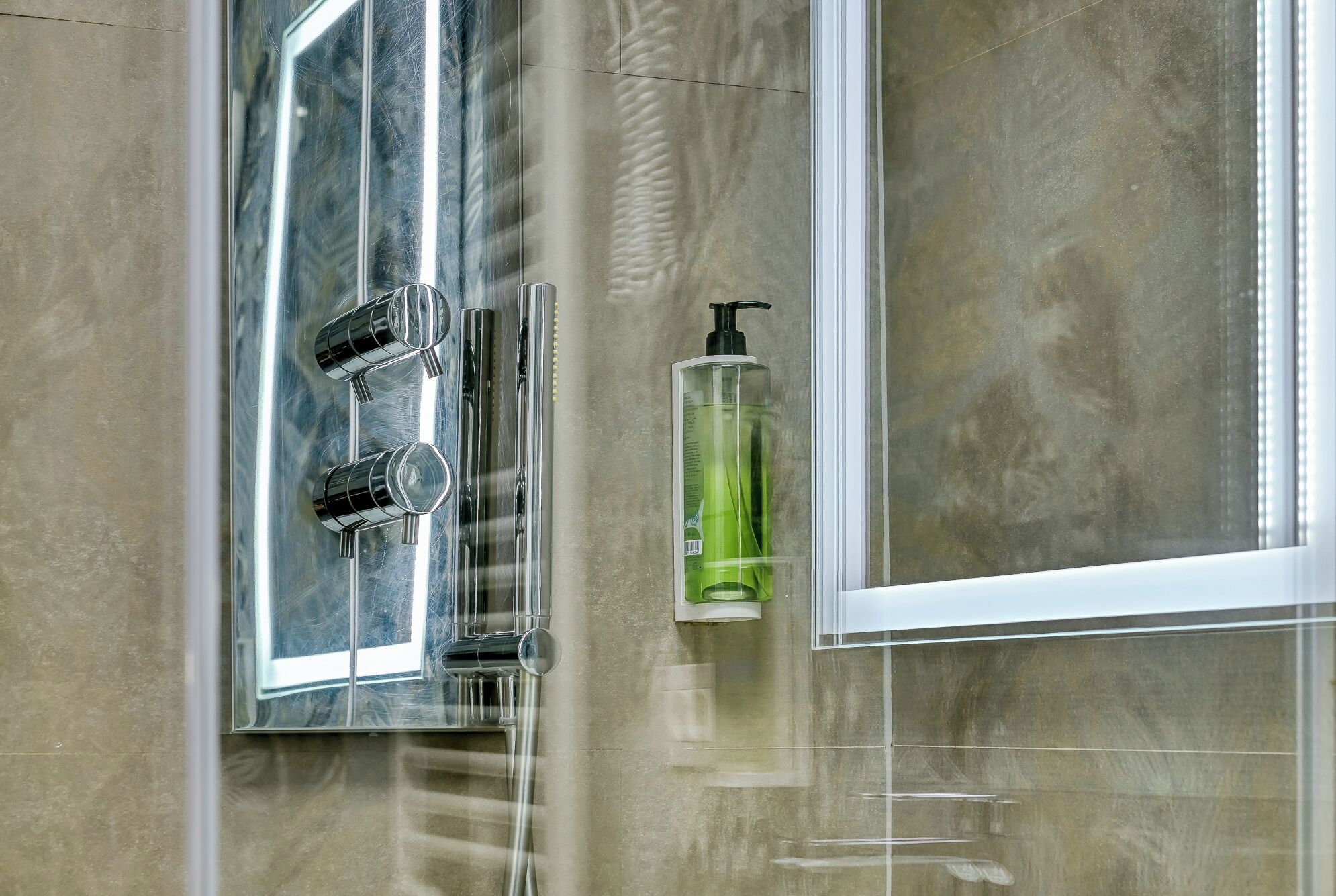 A bathroom with a glass shower enclosure, a handheld showerhead, and a green bottle of shampoo.
