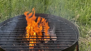 Un barbecue avec des flammes, entouré d’herbes hautes et d’une clôture, dans un cadre rural.
