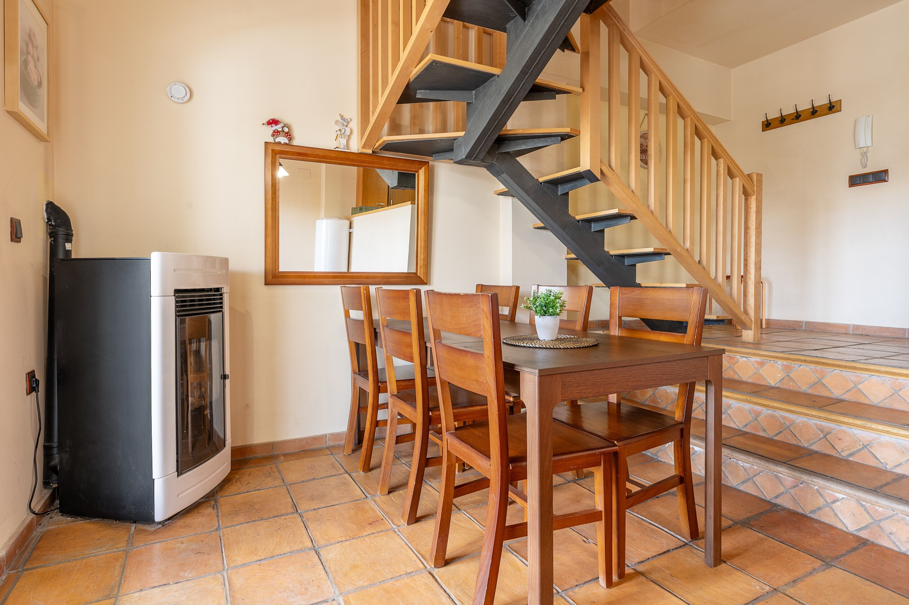 A dining area with a wooden table and chairs, a mirror, a potted plant, and a staircase with wooden railings.