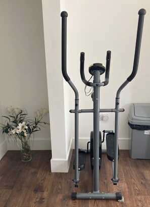 Een stilstaande hometrainer in een kamer met houten vloer, een prullenbak en een vaas met bloemen.