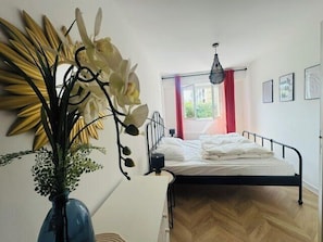 A bedroom with a bed, a window with curtains, a hanging light fixture, and a vase with flowers.