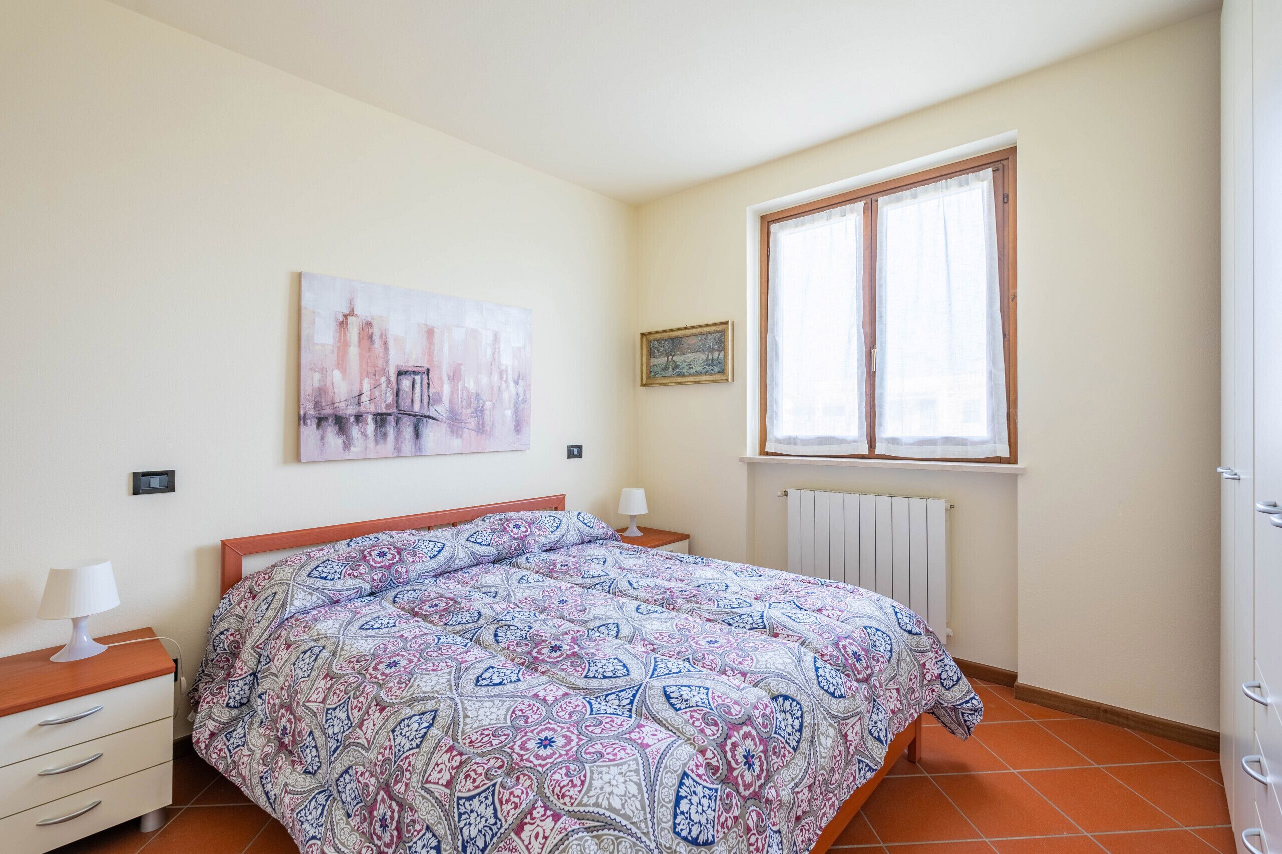 Ein Schlafzimmer mit Bett, Nachttisch, Kommode, einem Fenster mit Vorhängen und einer Heizung.