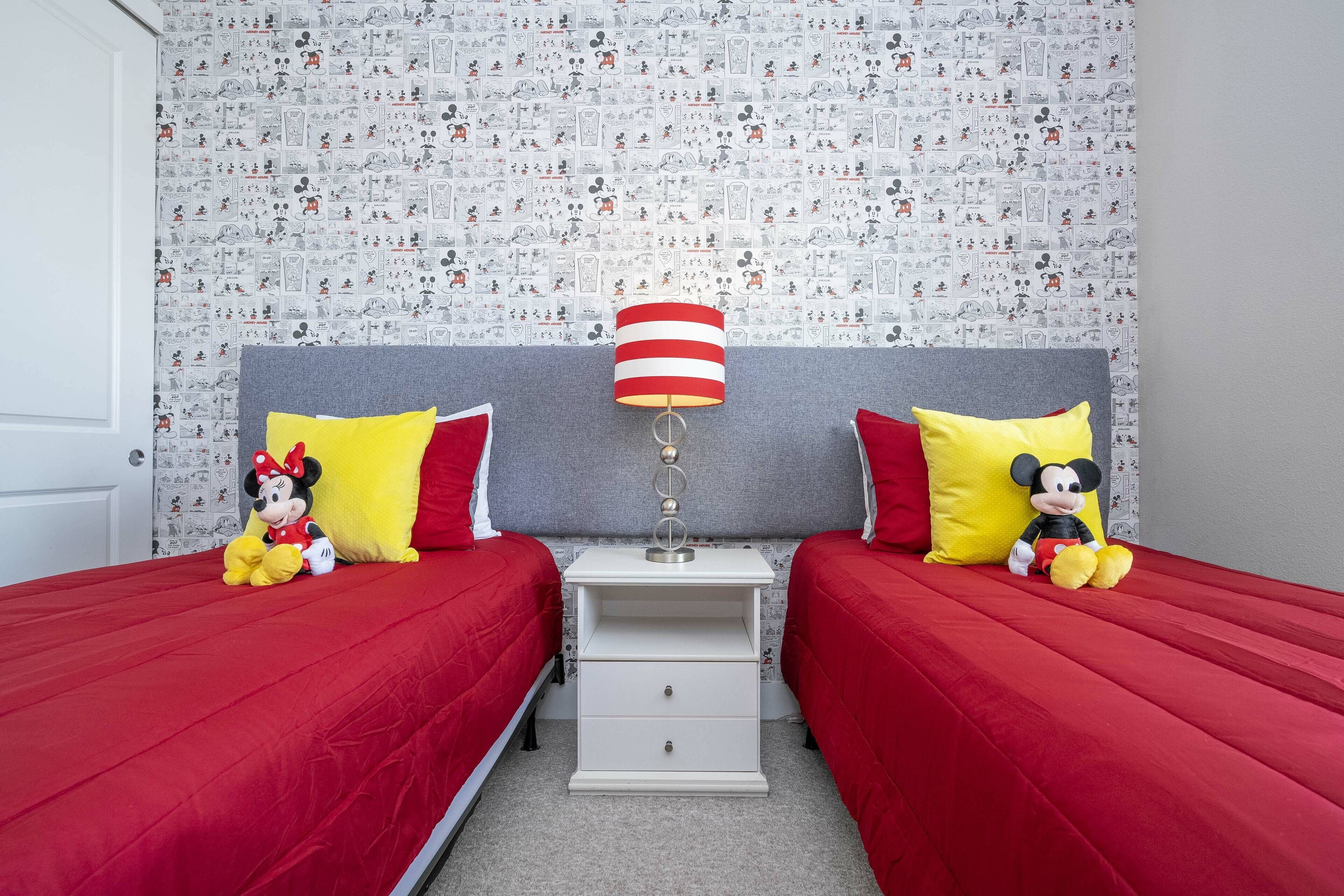 Un dormitorio con dos camas, cada una con ropa de cama roja y almohadas de Mickey Mouse. Entre las camas hay una mesita de noche blanca con una lámpara.