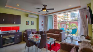 A modern living room with a dining area, kitchen, and a balcony with a view of buildings and greenery.