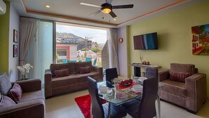 A living room with a glass dining table, two sofas, a ceiling fan, and a TV on the wall.