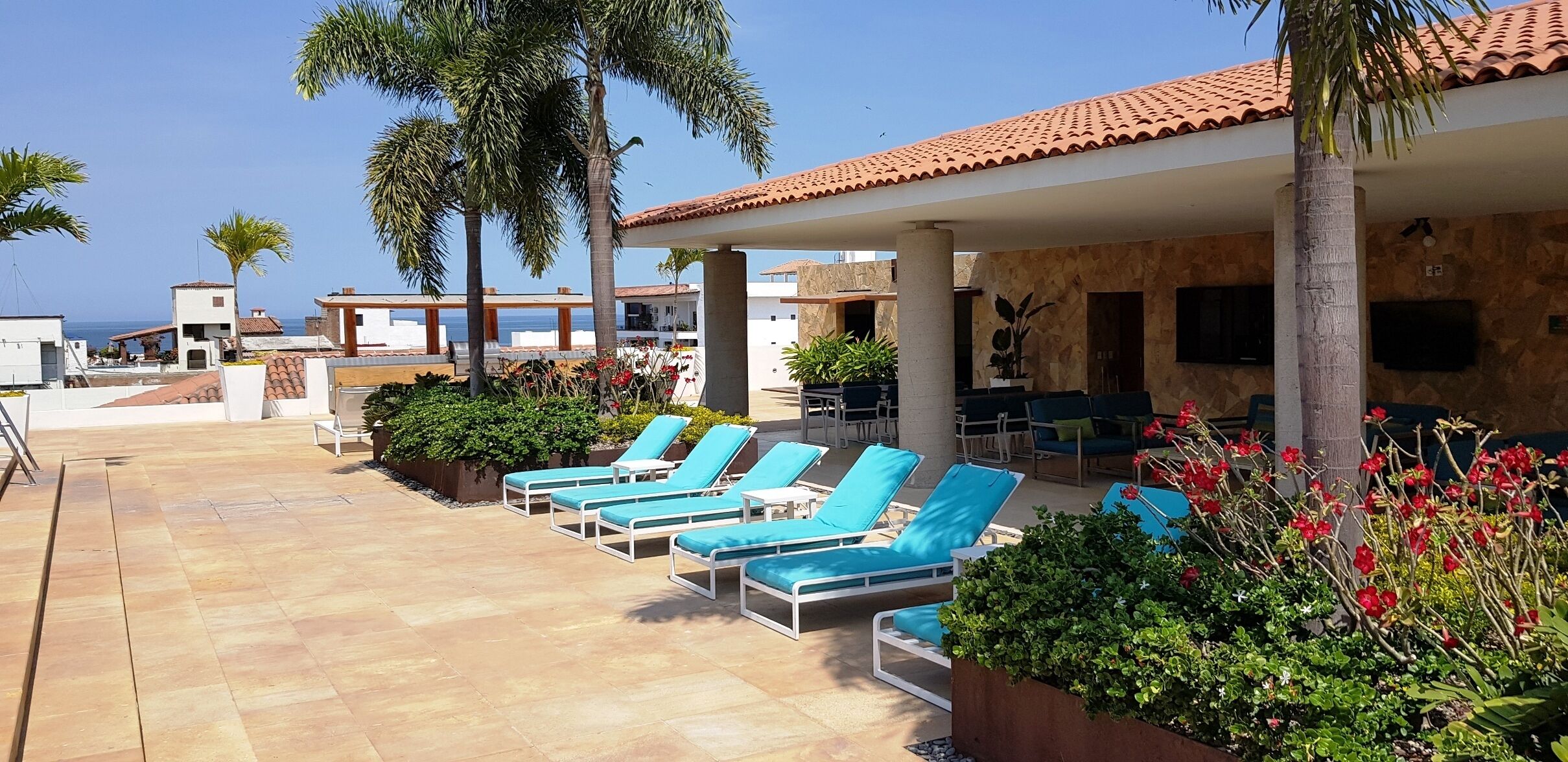Zona de piscina con tumbonas y palmeras, rodeada de edificios de estilo arquitectónico mediterráneo.