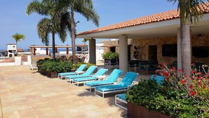 A pool area with sun loungers and palm trees, surrounded by buildings with a Mediterranean architectural style.