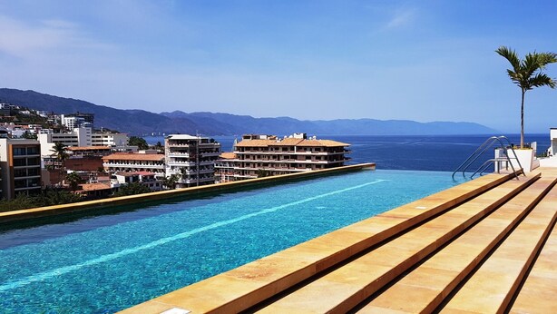 A swimming pool with a view of the sea and mountains, surrounded by buildings.