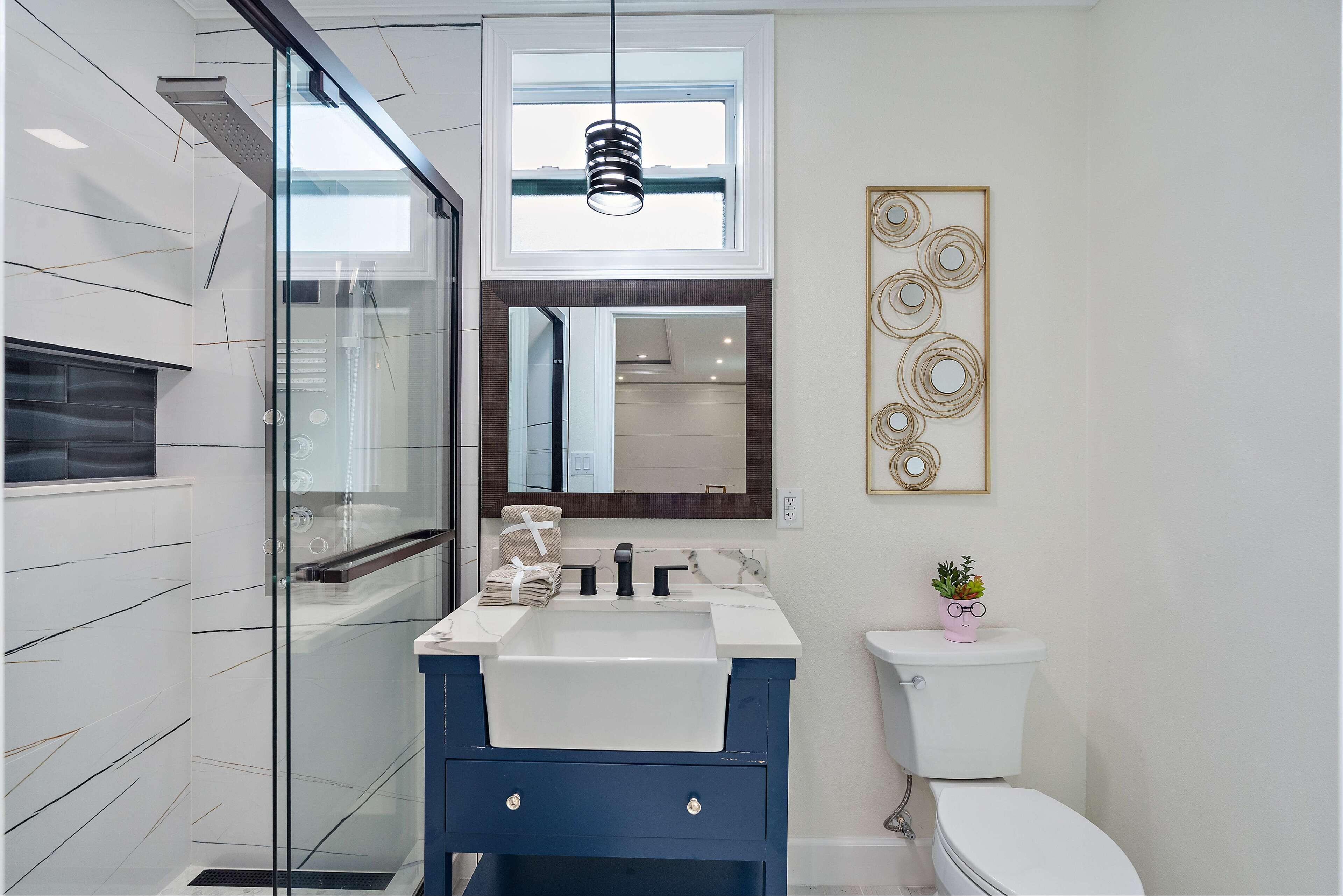 A modern bathroom with a glass shower enclosure, a white sink with a blue vanity, a toilet, and a decorative wall piece.