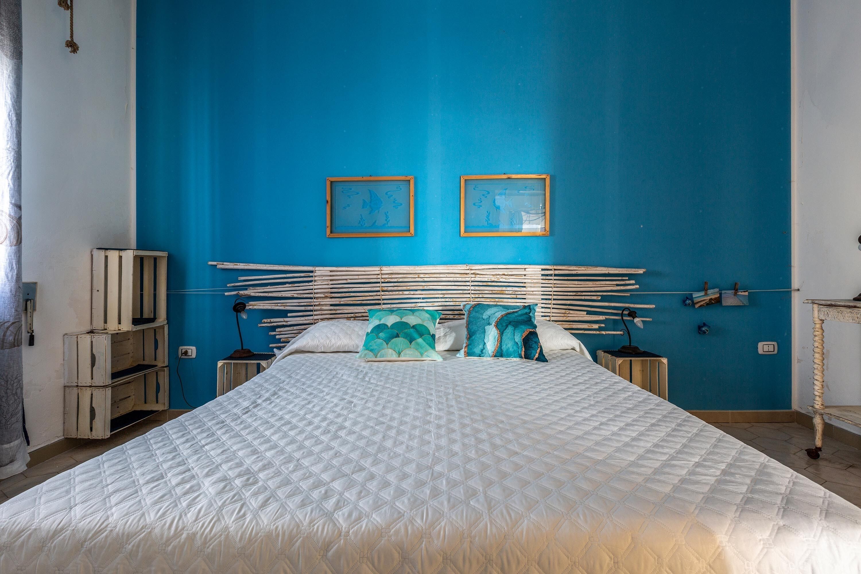 A bedroom with a large bed, two framed pictures on the wall, and a bedside table with a lamp.