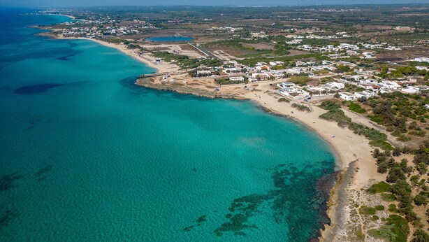 Vista aerea di una zona costiera con acque turchesi cristalline, una spiaggia sabbiosa e un gruppo di edifici.