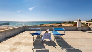 Una terrazza sul tetto con due poltrone blu, un tavolo con una bottiglia e un bicchiere, e vista mare e spiaggia.