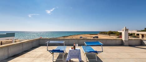 A rooftop terrace with two blue lounge chairs, a table with a bottle and glass, and a view of the sea and beach.