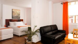 A hotel room with two beds, a black leather sofa, a wooden table, and a window with orange curtains. - Suites Machupicchu (Machu Picchu)