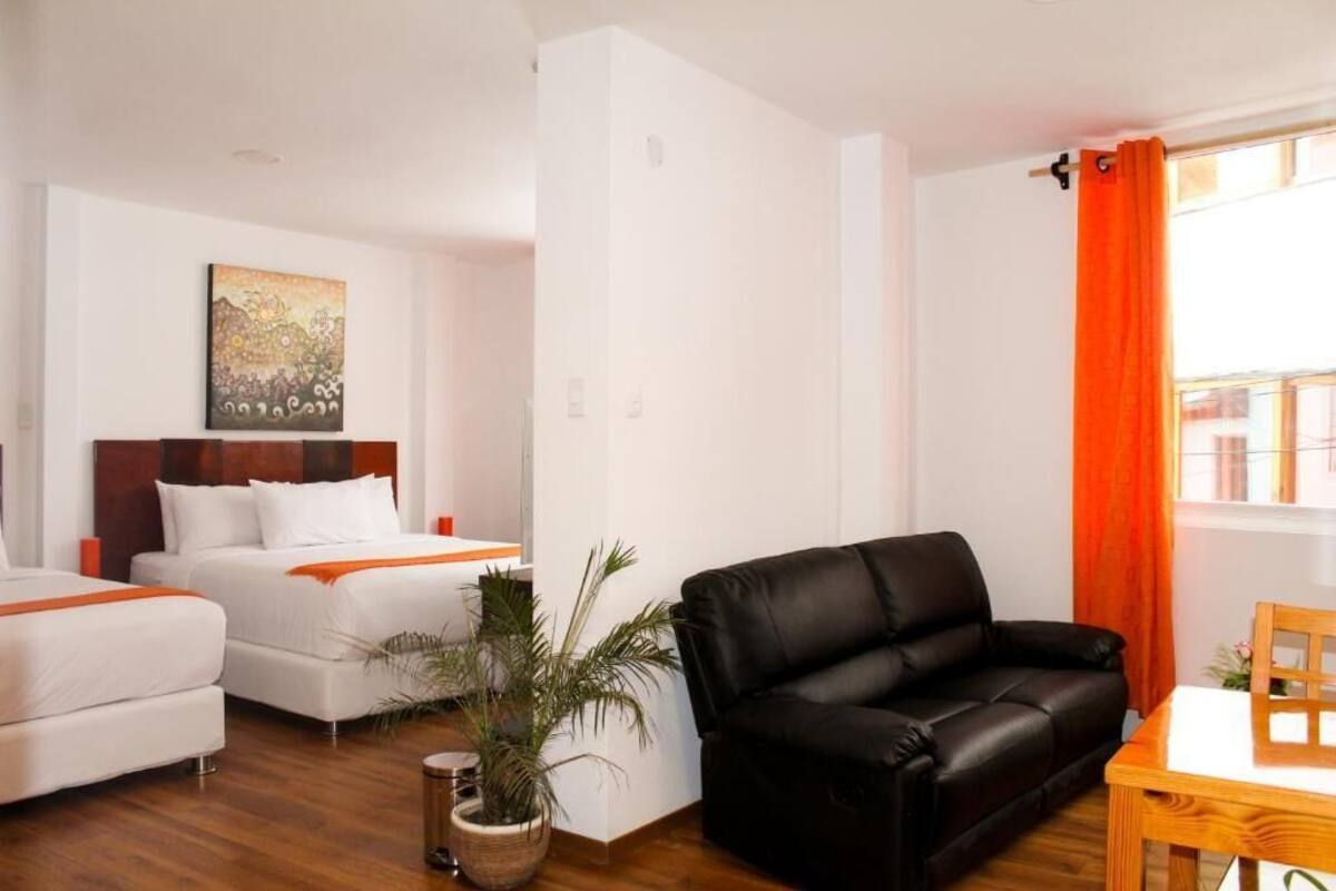 Quarto de hotel com duas camas, um sofá de couro preto, uma mesa de madeira e uma janela com cortinas laranja.