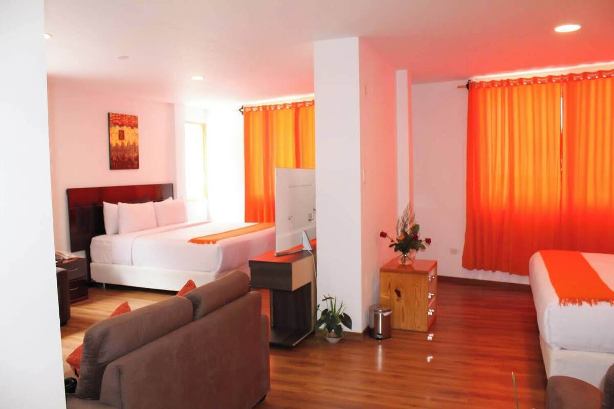 Quarto de hotel com cama, sofá, escrivaninha com computador e uma pequena planta.