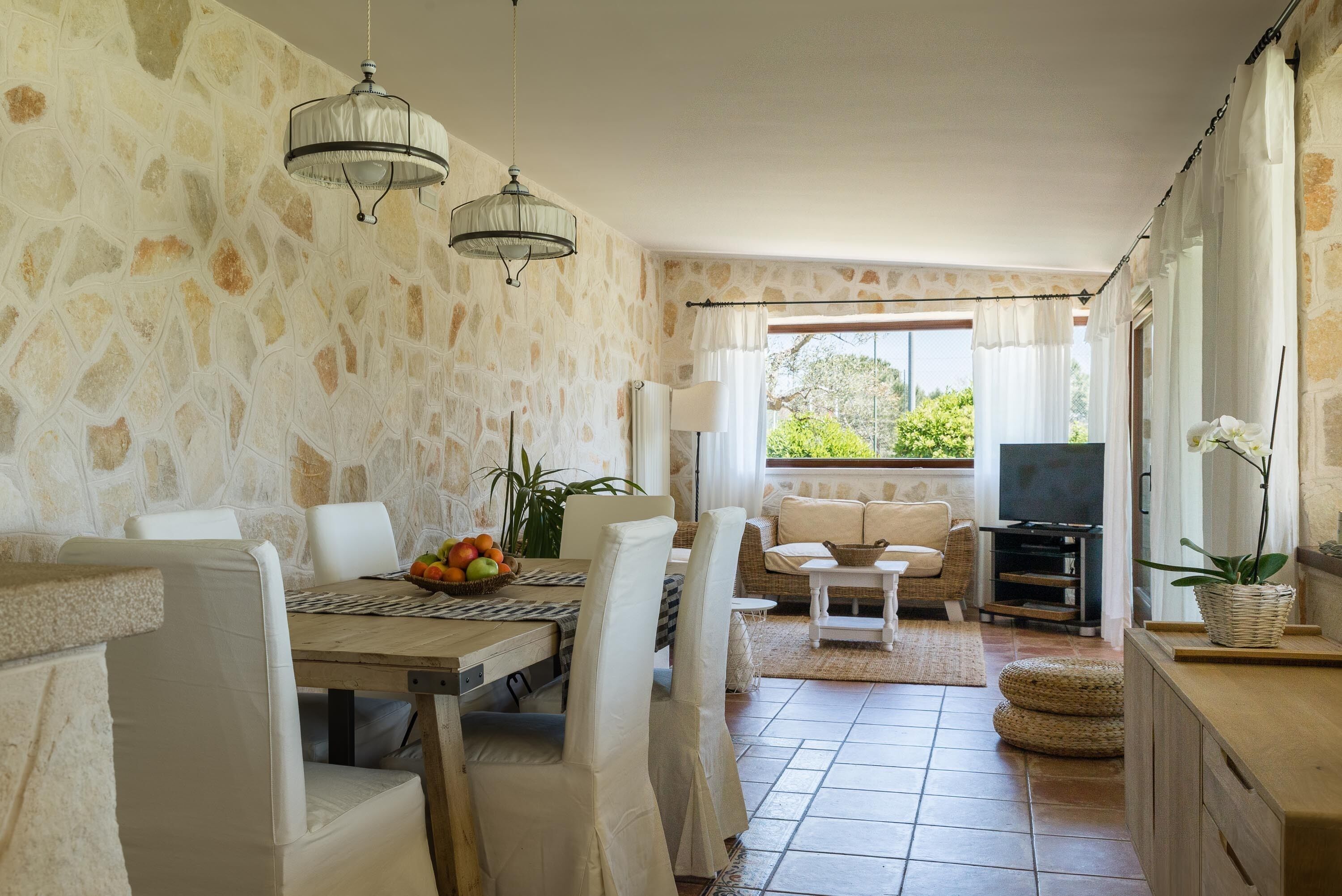 Un espace repas avec des murs en pierre, des meubles blancs, un lustre et une vue sur la verdure environnante.