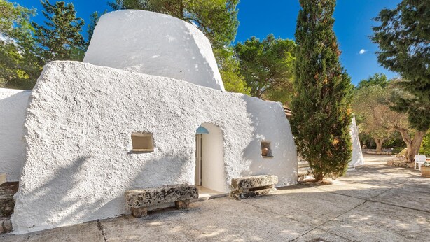 Un edificio blanco y rústico con techo plano, dos pequeñas puertas en arco y un banco de piedra al frente.