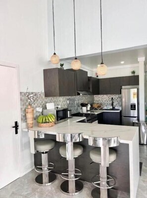 A modern kitchen with dark cabinets, a central island with bar stools, hanging pendant lights, and a tiled backsplash. - Tropical Garden Retreat in Valele, Ba - 2 Bedroom, 2 Bath Villa (Ba)
