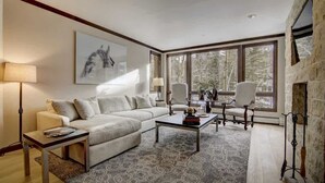 A modern living room with a sectional sofa, a coffee table, and a fireplace. There are large windows providing a view of the outdoors.