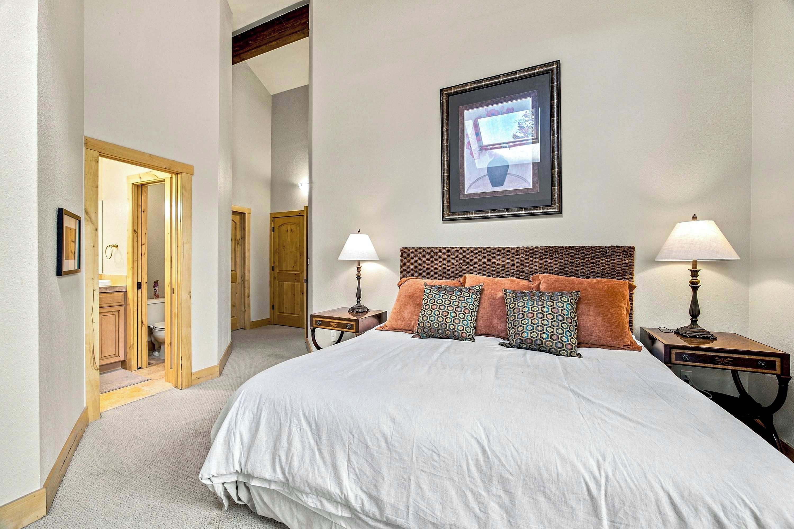 A hotel room with a large bed, two bedside tables with lamps, a framed picture on the wall, and a wooden door leading to a bathroom.