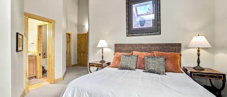 A hotel room with a large bed, two bedside tables with lamps, a framed picture on the wall, and a wooden door leading to a bathroom.