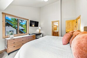 A bedroom with a large bed, a desk, a chair, a TV mounted on the wall, and a window with a view of a house and trees.