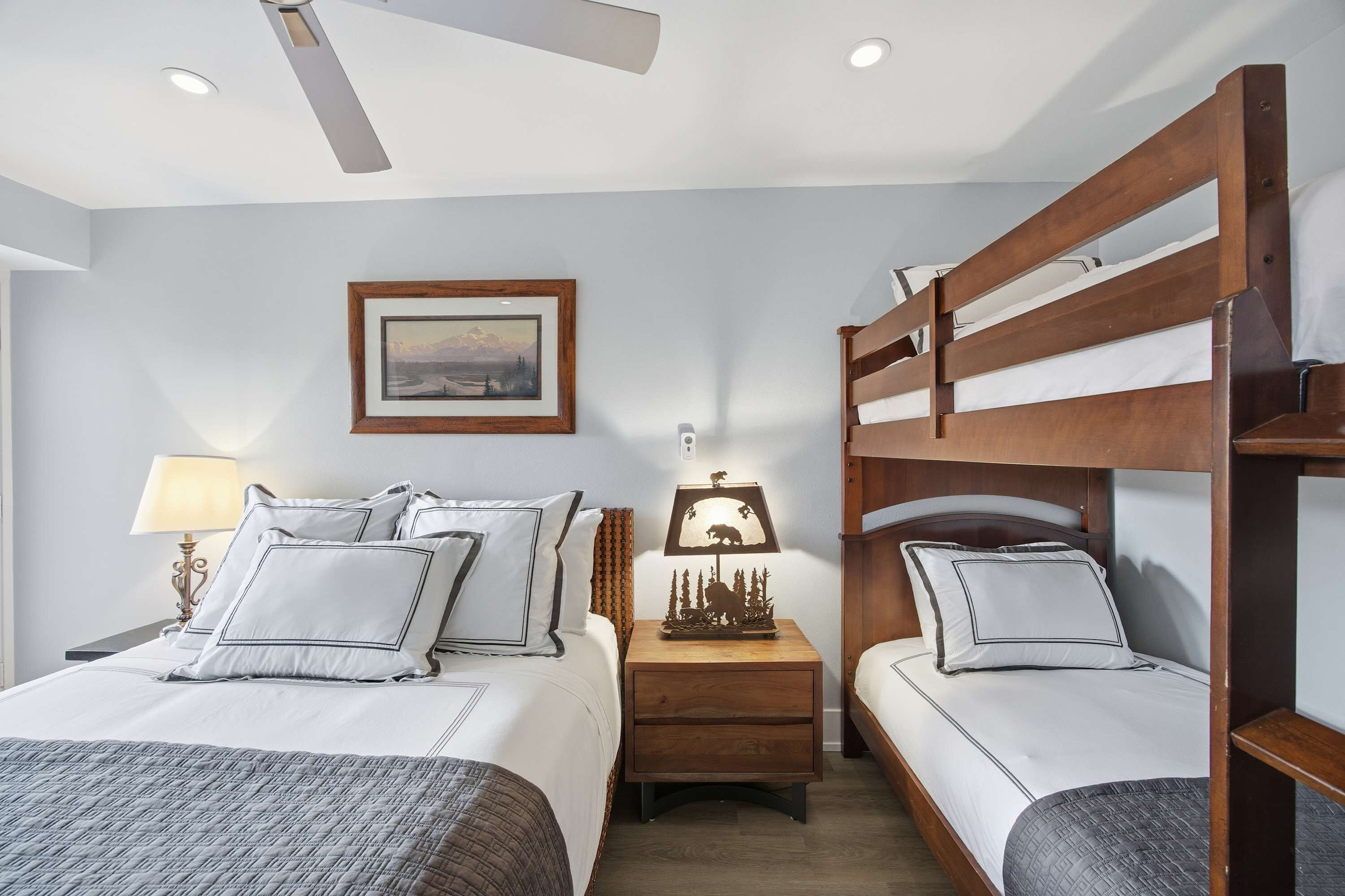 Une chambre d’hôtel avec un lit superposé, un lit simple, une table de chevet avec une lampe, un tableau encadré au mur et un ventilateur de plafond.