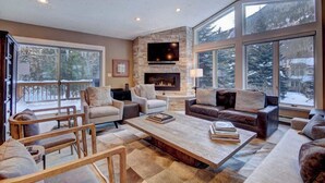 A cozy living room with a stone fireplace, comfortable seating, and large windows offering a view of a snowy landscape.