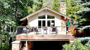 A two-story house with a stone chimney, a balcony with outdoor furniture, and a view of a forested hillside.