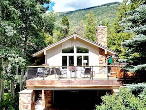 Una casa de dos pisos con chimenea de piedra, un balcón con mobiliario exterior y vista a una ladera boscosa.