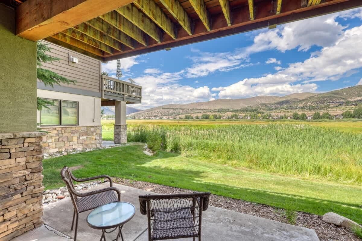 A covered patio with two wicker chairs and a glass table, overlooking a grassy field and distant hills.
