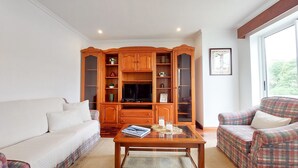 Un salon comprenant un canapé, un fauteuil à motifs, un meuble de rangement en bois avec des étagères, un téléviseur à écran plat et une table basse.