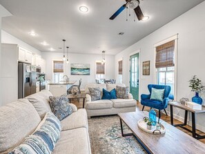A spacious living room with a sofa set, a coffee table, and a blue armchair. There is a kitchen in the background with a refrigerator and a dining area.