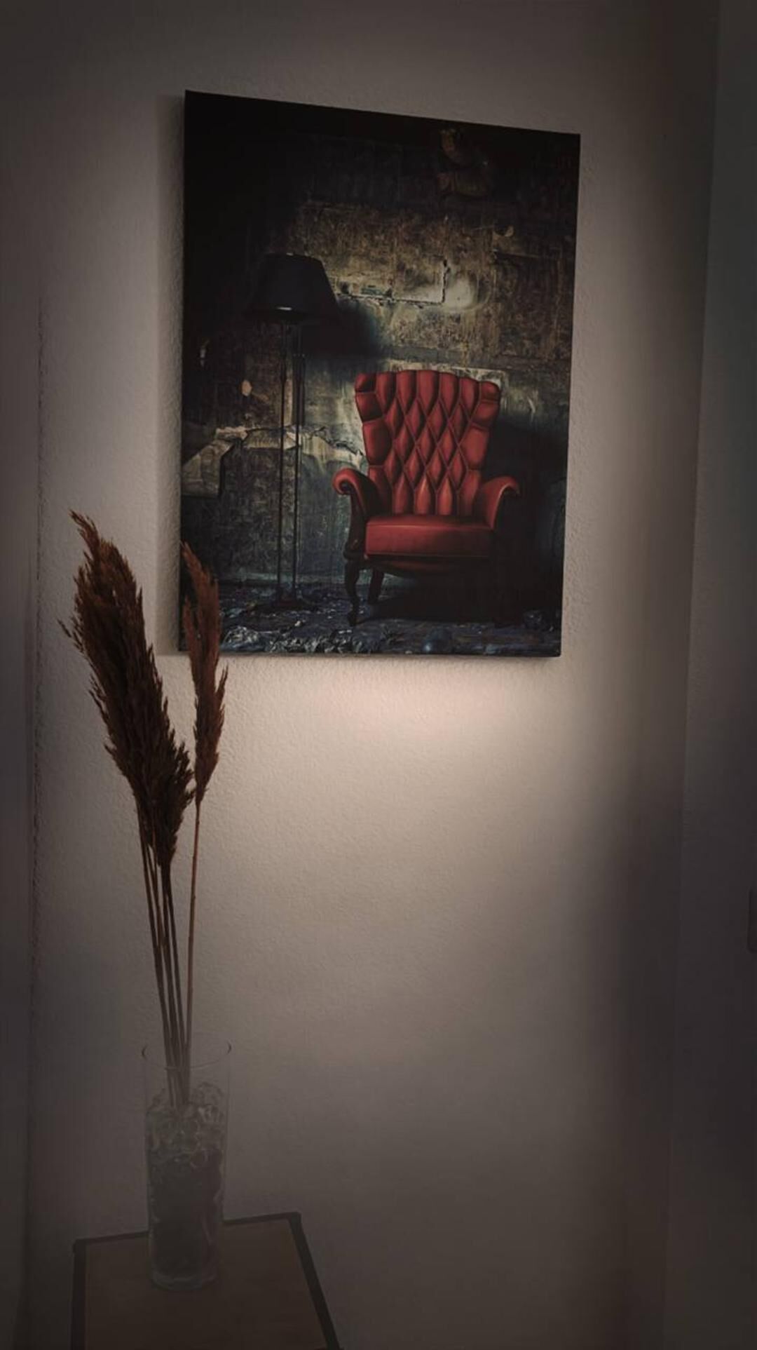 A framed artwork of a red chair against a textured wall, with a clear glass vase with dried plants on a table below.