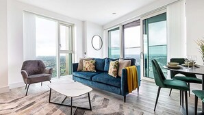 A modern living room with a blue sofa, a round coffee table, and a dining area with green chairs. - Modern Sky-High Flat In North Acton (London)