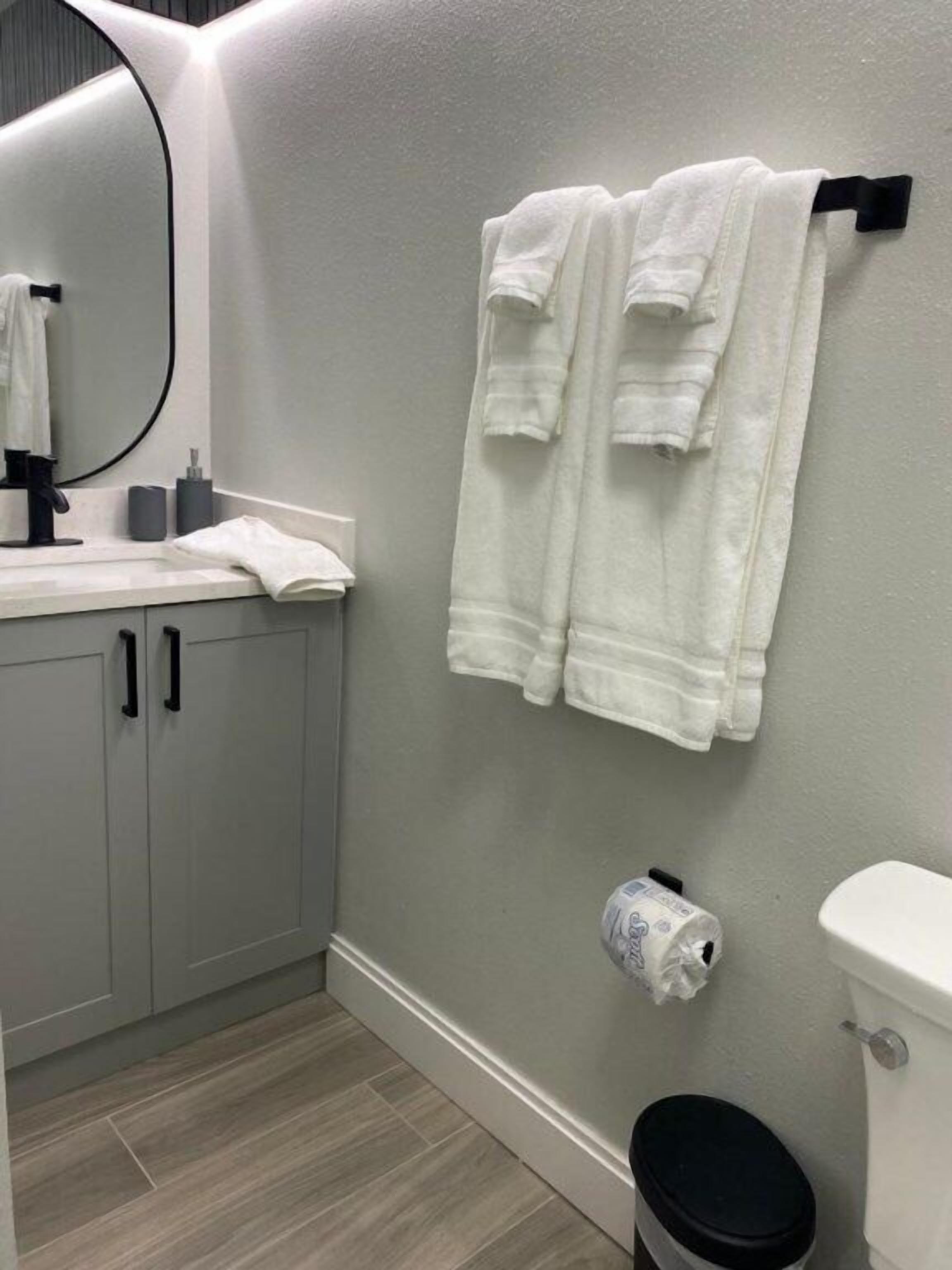Ein Badezimmer mit grauer Wand, einer weißen Toilette, einer schwarzen Mülltonne und einem Handtuchhalter mit zwei weißen Handtüchern.