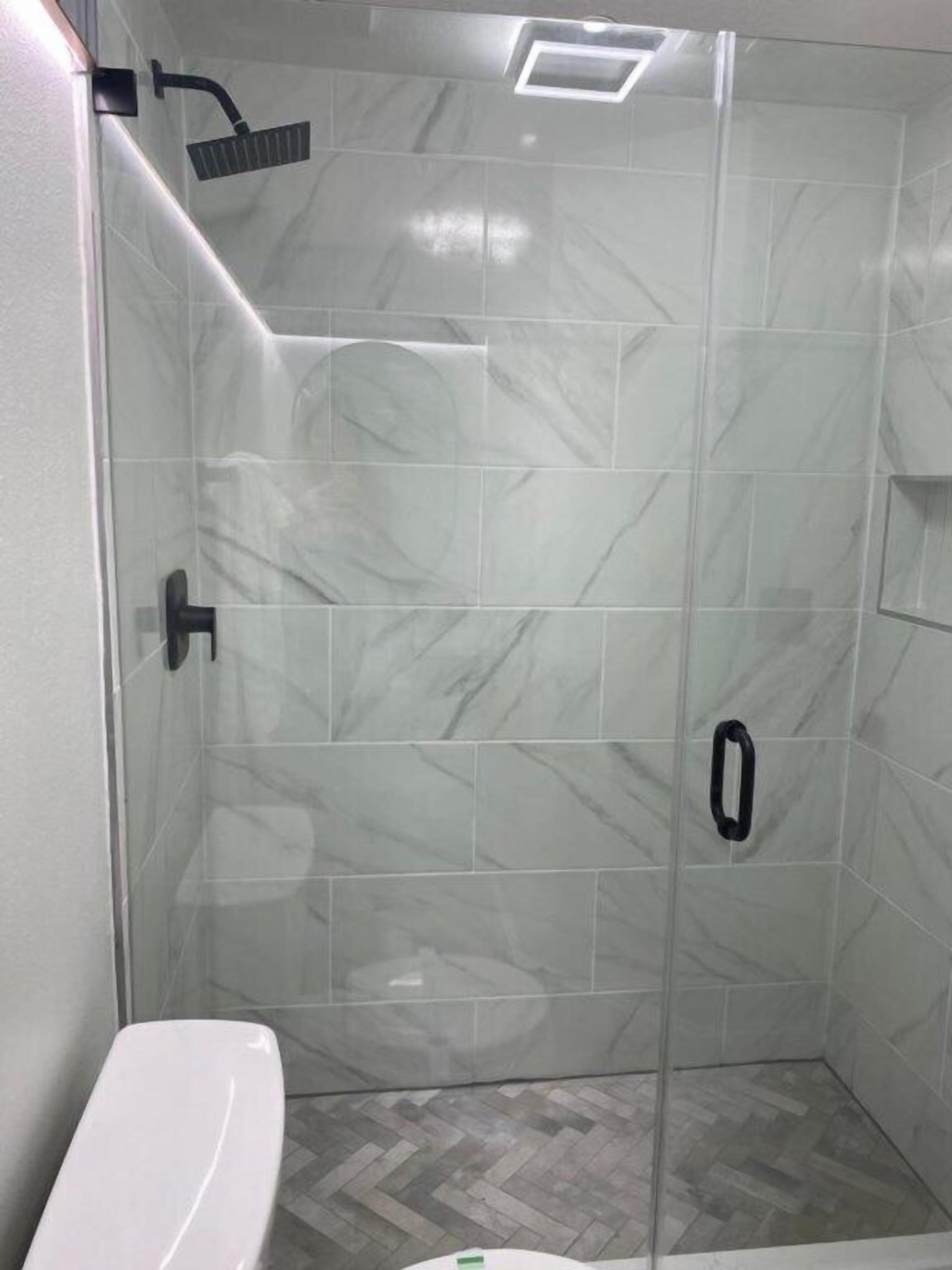 Un baño moderno con ducha envuelta en vidrio, azulejos de mármol blanco y suelo con motivo de espiga.
