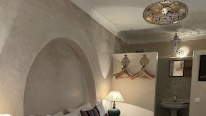 A neatly made bed with white linens, a striped pillow, and a rolled towel. There is a bedside table with a lamp and books. A hanging light fixture is above the bed. - Riad Alena Marrakech (Marrakech)
