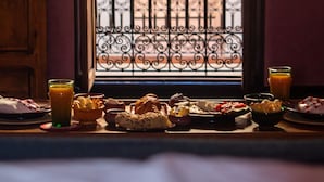 A breakfast table with various dishes and drinks, a window with decorative iron bars, and a view of a building outside. - Riad Alena Marrakech (Marrakech)