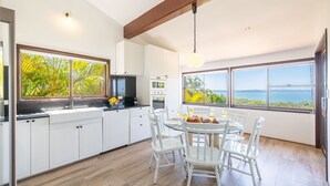 A modern kitchen with white cabinets, a dining area with white chairs, and large windows offering a view of the sea. - Casa Carinho, 24 Wallawa Rd - spectacular water views, pet friendly, air con and Wi-Fi (Nelson Bay)